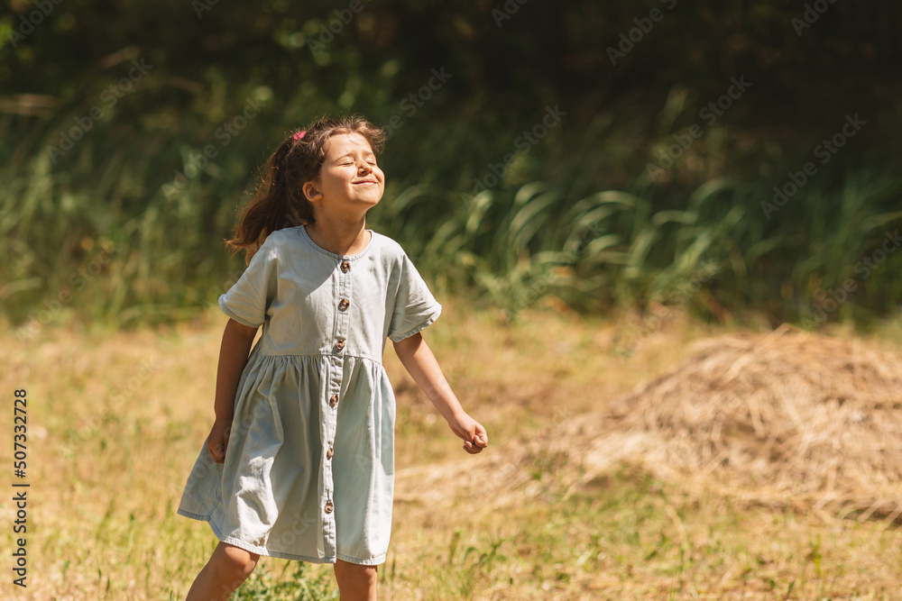 A little schoolgirl in a denim dress raised her head up and warms her nose in the sun, the child enjoys summer holidays, freedom and fun in the meadow A little schoolgirl in a denim dress raised her head up and warms her nose in the sun, the child enjoys summer holidays, freedom and fun in the meadow