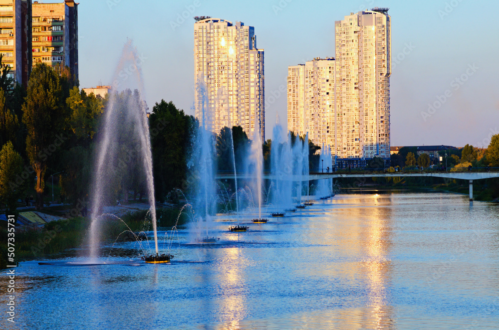 Obraz premium Picturesque sunset landscape of Rusanivka neighborhood at sunny day. Fountains in the city channel. Modern skyscrapers in the background. Landscape during the autumn season. Kyiv, Ukraine