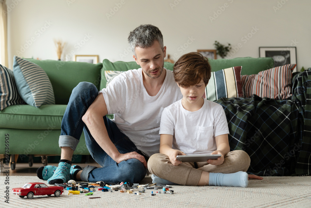 Dad and child sit together on the floor of the house and play constructor and video games on a tablet. Father and son spend their free time together. Dad and child sit together on the floor of the house and play constructor and video games on a tablet. Father and son spend their free time together.