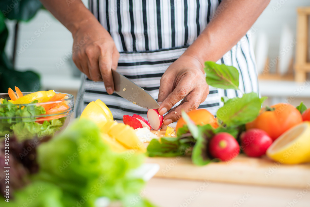 Close-up of chef's hand using stainless steel knife to cut radishes on a wooden cutting board and vegetables such as tomatoes, lettuce and bell peppers prepared on the salad table in the kitchen.
