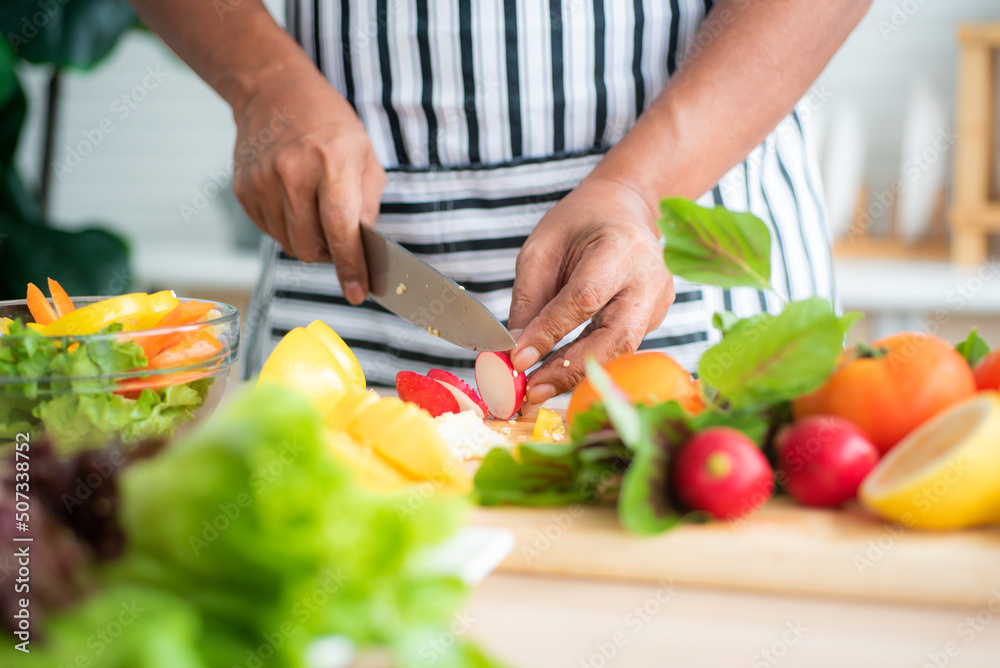 Close-up of chef's hand using stainless steel knife to cut radishes on a wooden cutting board and vegetables such as tomatoes, lettuce and bell peppers prepared on the salad table in the kitchen. Close-up of chef's hand using stainless steel knife to cut radishes on a wooden cutting board and vegetables such as tomatoes, lettuce and bell peppers prepared on the salad table in the kitchen.