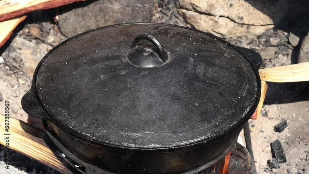 A cast-iron cauldron is closed with a lid and stands on a fire. Cooking outdoors in national utensils. High quality 4k footage.
