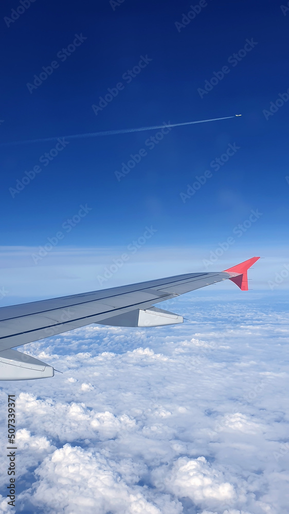 Soft clouds cover the sky during the flight. View of the wing of an airplane flying above the clouds at high altitude under a blue sky from the passenger window Soft clouds cover the sky during the flight. View of the wing of an airplane flying above the clouds at high altitude under a blue sky from the passenger window