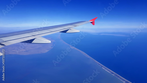 Soft clouds cover the sky during the flight. View of the wing of an airplane flying above the clouds at high altitude under a blue sky from the passenger window