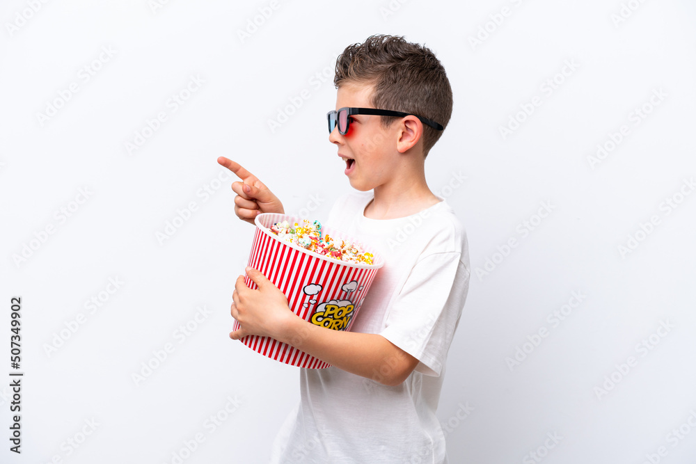 Little caucasian boy isolated on white background with 3d glasses and holding a big bucket of popcorns while pointing front
