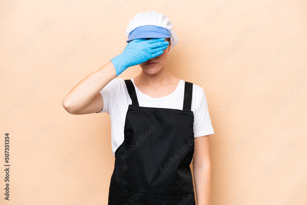 Fishmonger caucasian woman wearing an apron and holding a raw fish isolated on beige background covering eyes by hands. Do not want to see something