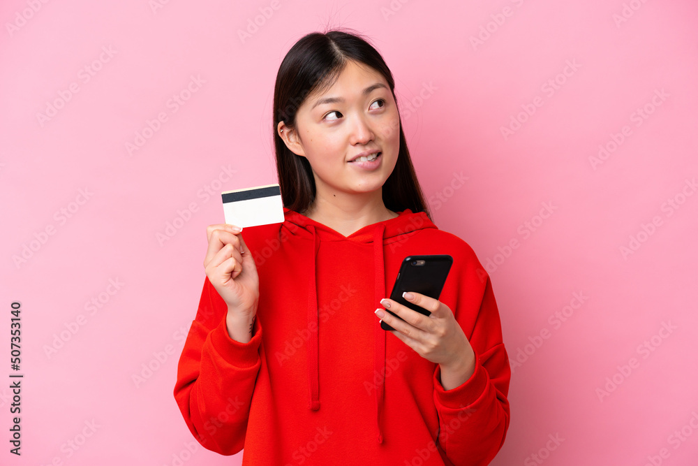 Young Chinese woman isolated on pink background buying with the mobile with a credit card while thinking