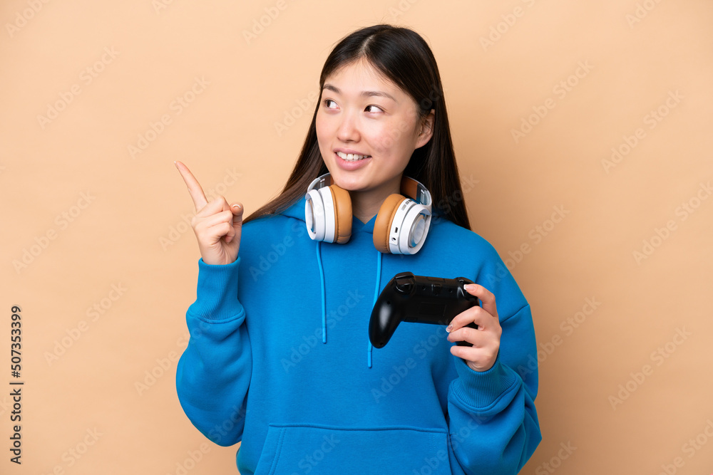 Young Chinese man playing with a video game controller isolated on beige background pointing up a great idea Young Chinese man playing with a video game controller isolated on beige background pointing up a great idea