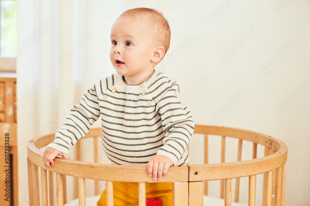A handsome little boy in a striped blouse is holding on to the sides of the crib. A handsome little boy in a striped blouse is holding on to the sides of the crib.