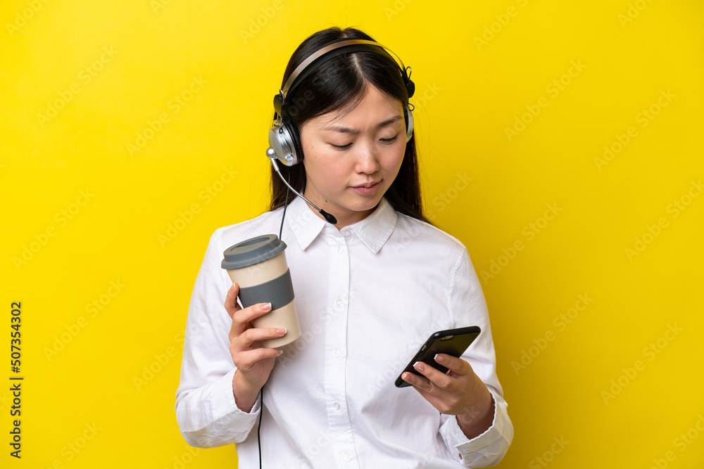 Telemarketer Chinese woman working with a headset isolated on yellow background holding coffee to take away and a mobile