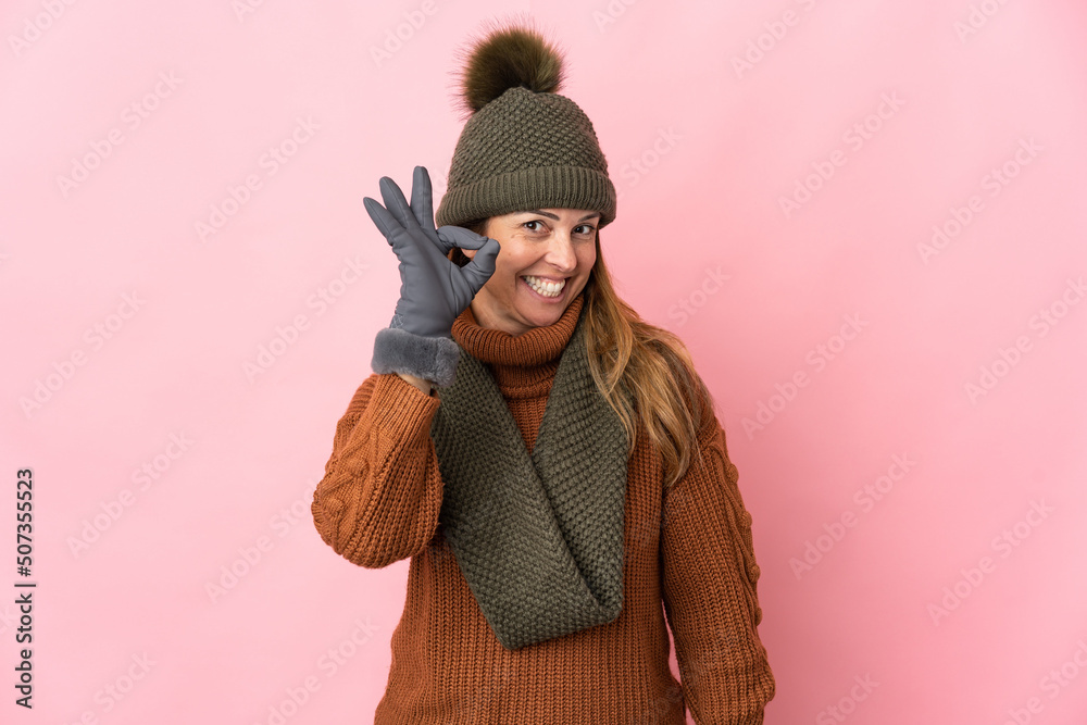 Middle age woman with winter hat isolated on pink background showing ok sign with fingers Middle age woman with winter hat isolated on pink background showing ok sign with fingers