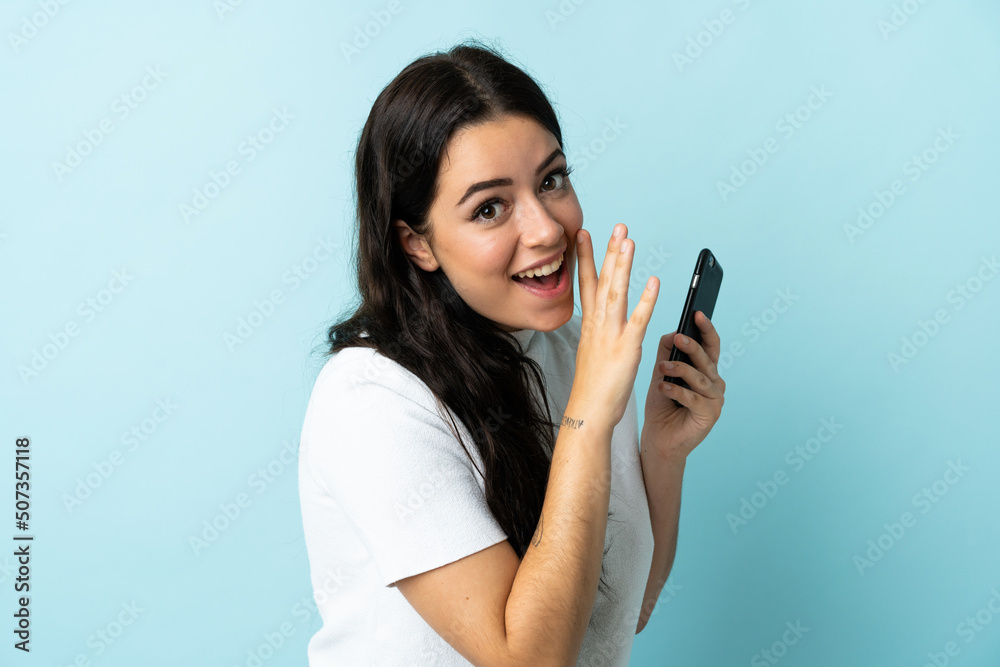 Young woman using mobile phone isolated on blue background whispering something Young woman using mobile phone isolated on blue background whispering something