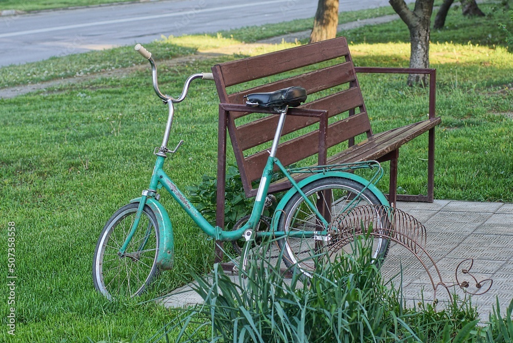one old bicycle stands in the green grass near the brown bench on the street in the park