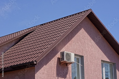 attic of a private pink house with windows and a white air conditioner on a wall under a brown tiled roof against a blue sky
