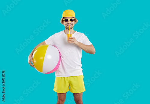 Summer rest. Happy male beach vacationer enjoys freshly squeezed orange juice isolated on light blue background. Smiling man in summer clothes and with inflatable beach ball drinks juice through straw photo