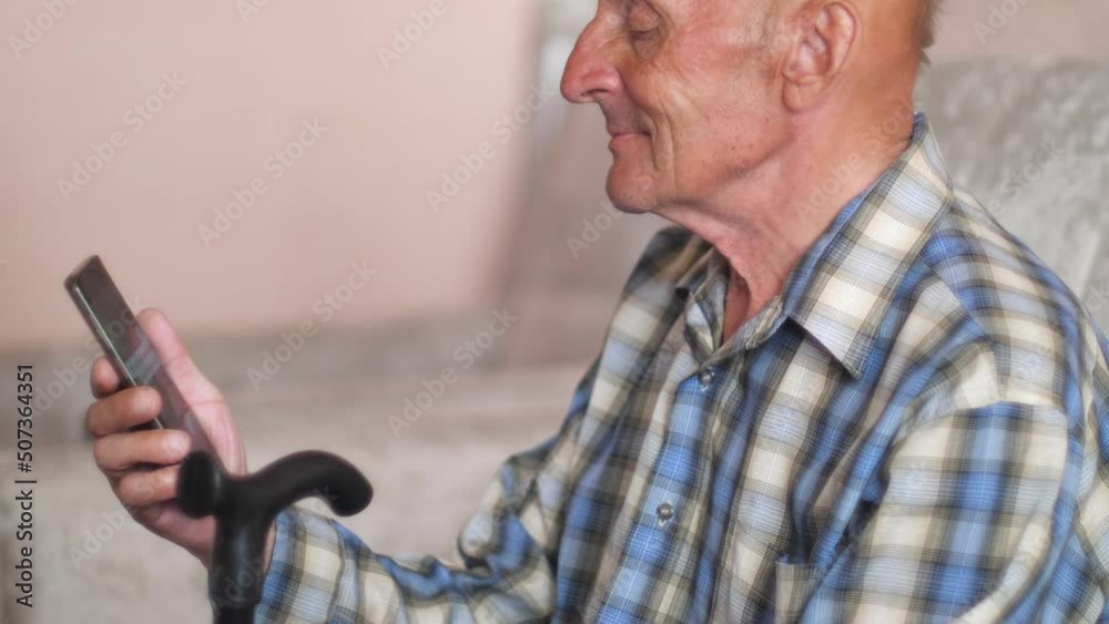 caucasian peppy pensioner with a smartphone and a walking stick sits on the couch at home.Smiling elderly man 70-80 years old spends leisure time with a phone, social networks caucasian peppy pensioner with a smartphone and a walking stick sits on the couch at home.Smiling elderly man 70-80 years old spends leisure time with a phone, social networks