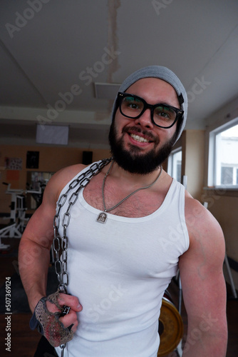 Portrait of a young man in a gray hat, white tank top with glasses and a beard in an old school gym. A guy in sports gloves and an iron chain on his shoulder. Sport and healthy lifestyle concept
