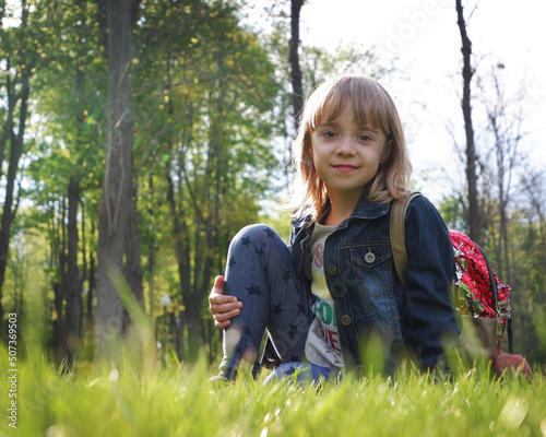 Smiling teenage girl in a stylish denim jacket and with a backpack in the park posing outdoors. A child with blond hair. Schoolchild concept