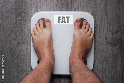 Top view of the feet of a barefoot man standing on smart scales, on whose balance it is written fat
