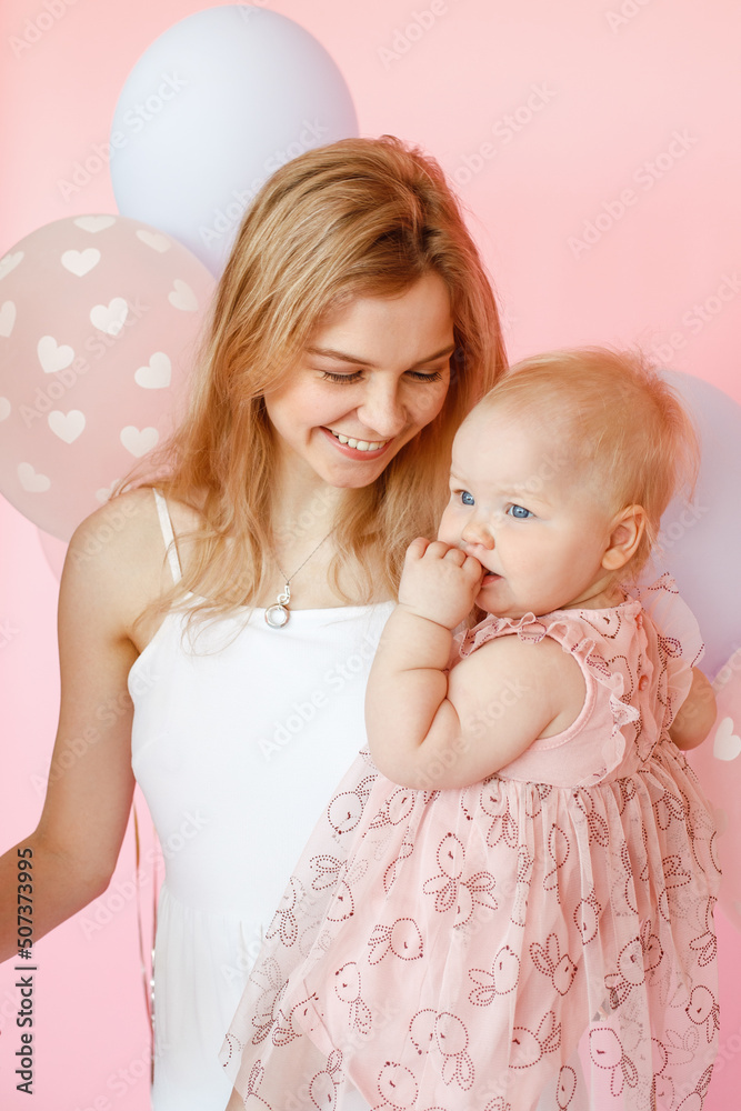 Portrait closeup of young smiling mother holding cute baby girl in holiday dress on pink background. First birthday party, pink decor and balloons. Happy moments and family holiday celebration Portrait closeup of young smiling mother holding cute baby girl in holiday dress on pink background. First birthday party, pink decor and balloons. Happy moments and family holiday celebration