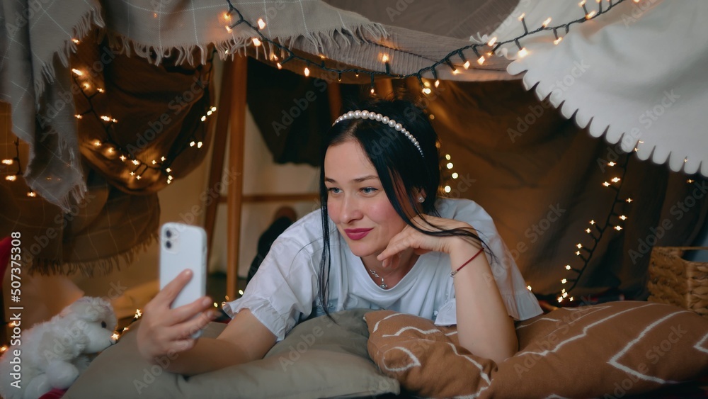 A young woman takes a selfie on her smartphone while lying in a tent of blankets and pillows in the bedroom. Leisure A young woman takes a selfie on her smartphone while lying in a tent of blankets and pillows in the bedroom. Leisure