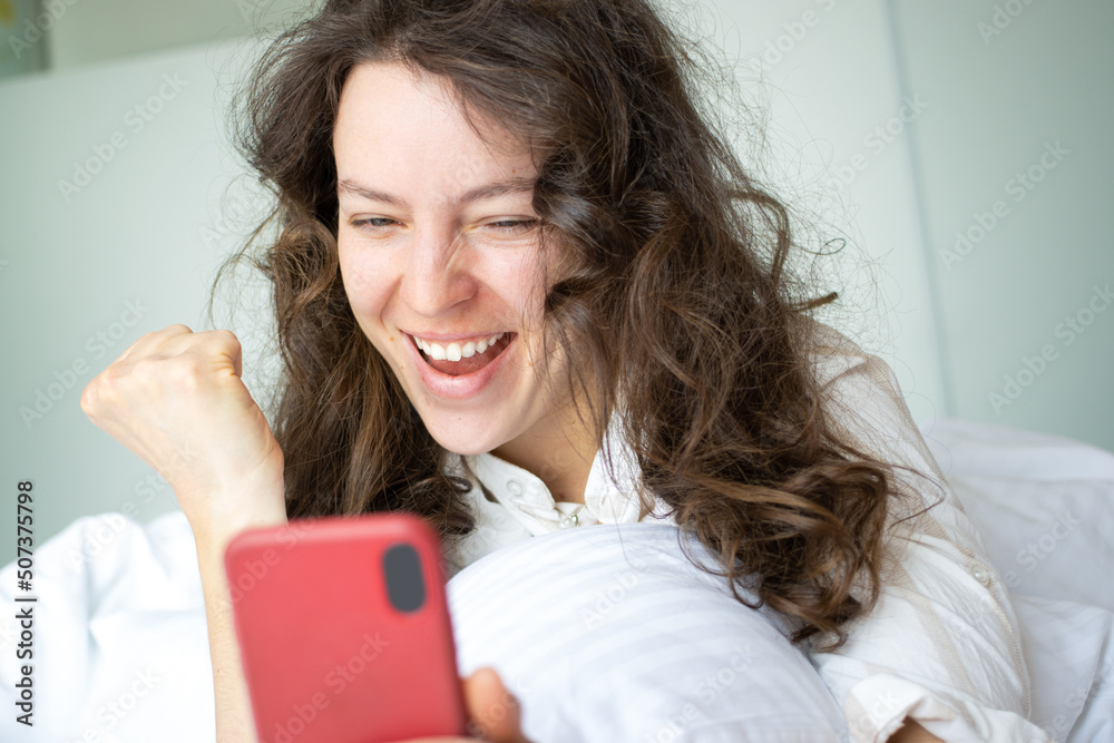 Excited young curly woman doing winner gesture, looking at mobile phone lying in white bed indoors. Feeling euphoria. Celebrate online success victory, new opportunity, got new job, great result Excited young curly woman doing winner gesture, looking at mobile phone lying in white bed indoors. Feeling euphoria. Celebrate online success victory, new opportunity, got new job, great result