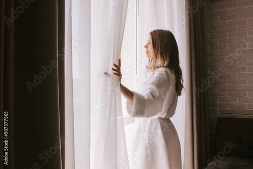 A young caucasian woman in a white robe opens the curtains meeting the dawn looking out the window. Morning of the bride
