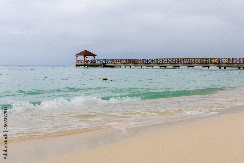 Fototapeta premium A pier goes into the sea,at the end there is a gazebo.The blue sea water and the sky are separated by the horizon line,a pier.Caribbean Coast in the Dominican Republic.A wave runs over the sandy shore