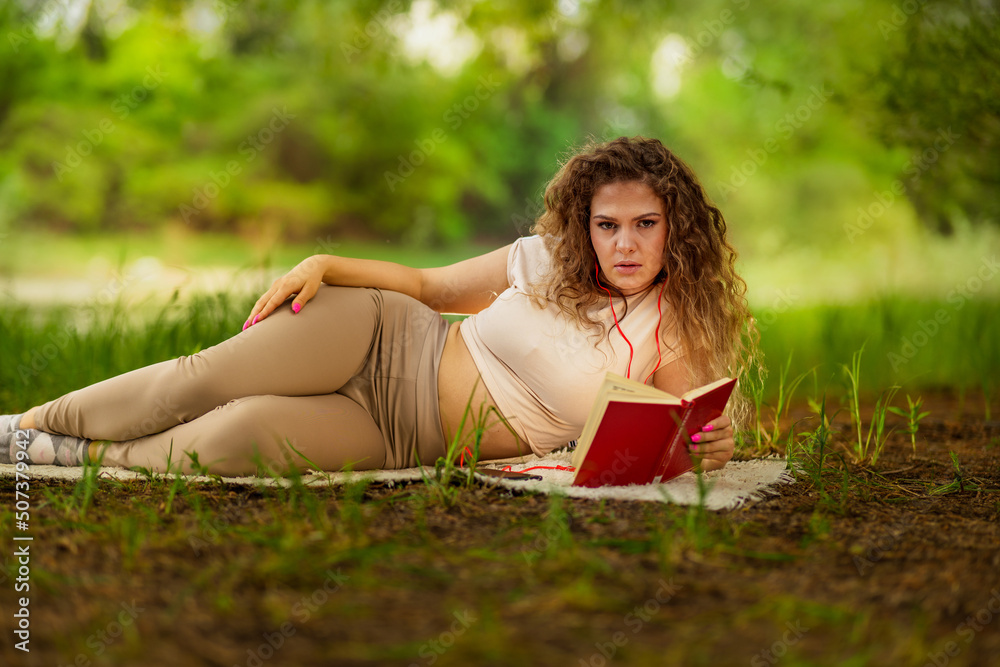 A beautiful young woman with laid hair lying on a blanket in the grass while reading a book and listening to music. A beautiful young woman with laid hair lying on a blanket in the grass while reading a book and listening to music.