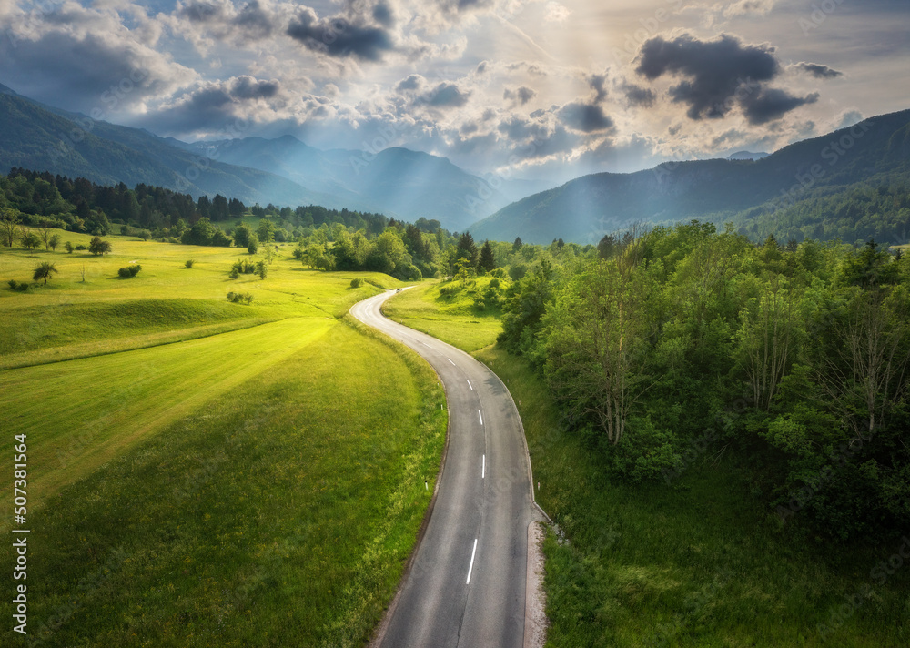 Aerial view of road in green meadows at sunset in summer. Top view from drone of rural road, mountains, forest. Beautiful landscape with roadway, sun rays, trees, hills, green grass, clouds. Slovenia - obrazy, fototapety, plakaty 