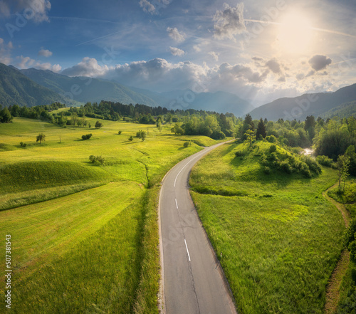 Aerial view of road in green meadows and hils at sunset in summer. Top view from drone of rural road, mountains, forest. Beautiful landscape with roadway, trees, green grass, sky with clouds. Slovenia