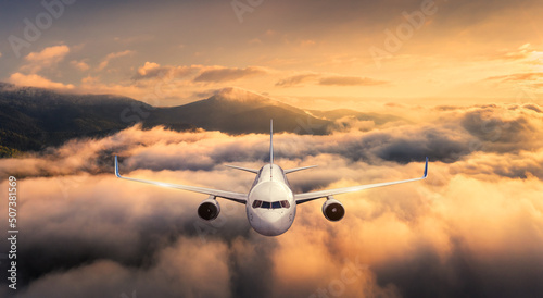 Airplane is flying above the clouds at sunset in summer. Landscape with passenger airplane, mountains, orange sky. Aircraft is taking off. Business travel. Commercial plane. Aerial view. Transport