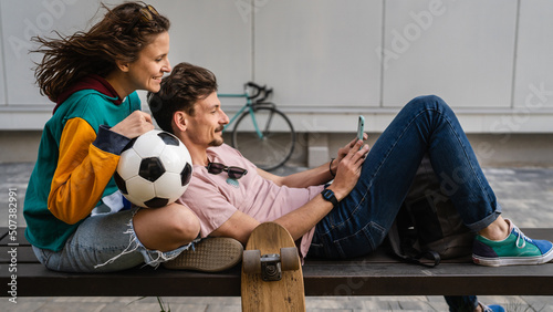 Couple woman and young caucasian man modern happy adult male and female boyfriend and girlfriend smile sitting in front of stadium holding soccer ball outdoor in day football game concept copy space photo