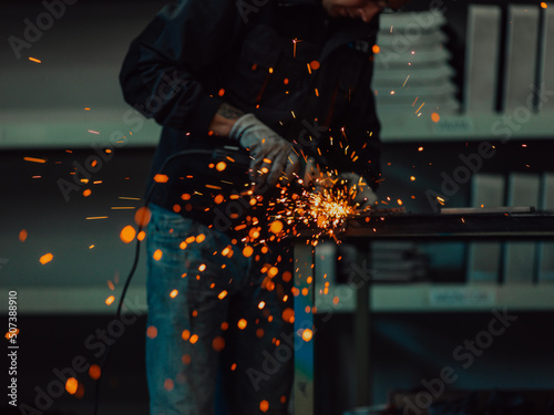 Fototapeta Naklejka Na Ścianę i Meble -  Heavy Industry Engineering Factory Interior with Industrial Worker Using Angle Grinder and Cutting a Metal Tube. Contractor in Safety Uniform and Hard Hat Manufacturing Metal Structures.