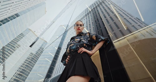 Wide angle urban style seductive girl wearing short skirt colored jacket cool futuristic glasses looks into camera standing by glass wall high skyscraper modern building. Handsome female fashion model photo