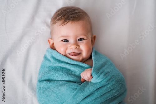 Top view of happy little cheeky 6 month old baby wrapped naked in turquoise towel and smiles & sticks tongue out at camera on a white background

