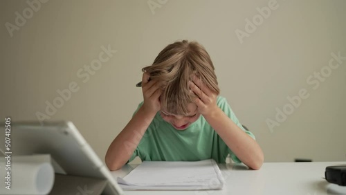 7 yeas old boy sitting at table in room and crying while working on hard homework. Frustrated young boy lays head done on schoolwork. photo