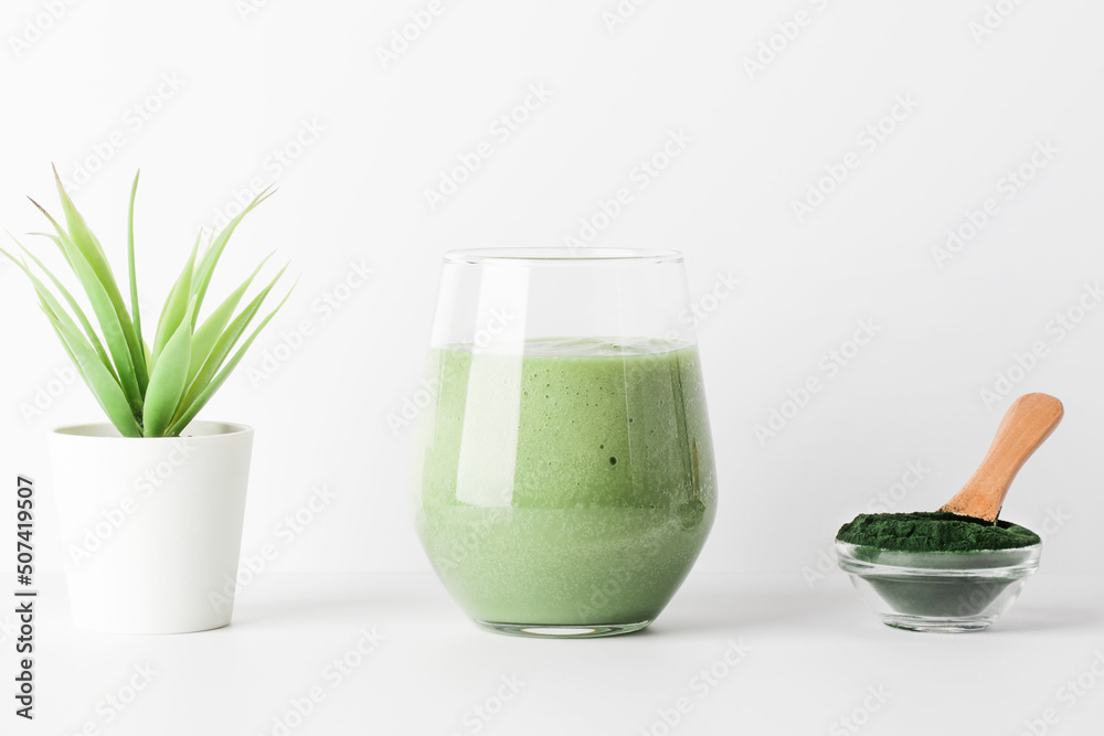 green smoothie in glass and spirulina algae powder in bowl with spoon, flower on white background. self care and healthy lifestyle concept.