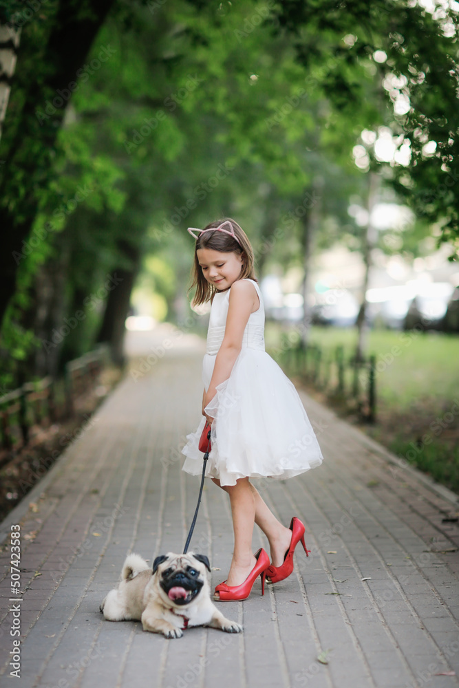 European girl child in white dress in red mother's shoes with heels on walk with pug, joyful child in summer with pet European girl child in white dress in red mother's shoes with heels on walk with pug, joyful child in summer with pet