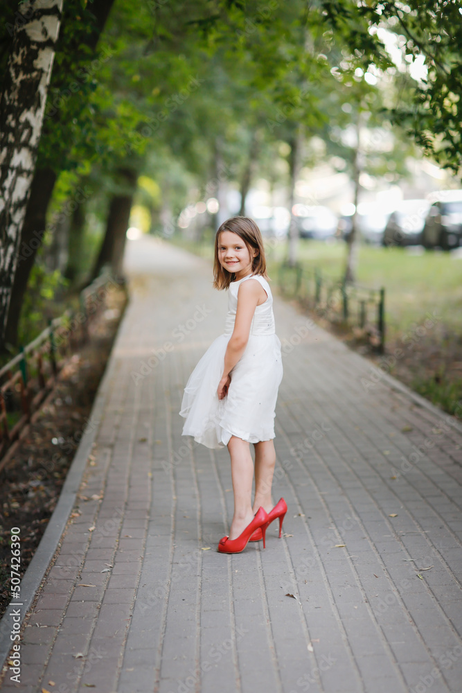Happy stylish baby girl in a white dress and adult high-heeled shoes jumps and runs along the sidewalk in summer. Happy stylish baby girl in a white dress and adult high-heeled shoes jumps and runs along the sidewalk in summer.