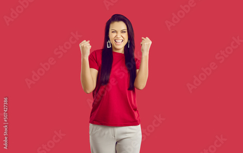 Joyful excited woman clenching her fists rejoicing in her victory or success on red background. Happy caucasian woman laughing shouting yes and happily clenching fists looking at camera. Banner.