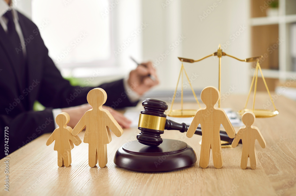 Gavel, sound block and little wooden figures of parents and children placed on desk in courthouse up close, judge and scales of justice in background. Family law, court, divorce, child custody concept - obrazy, fototapety, plakaty 