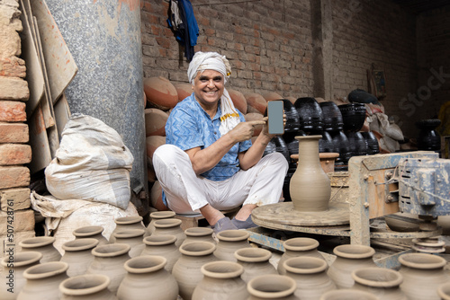 Photography Portrait of old man showing blank screen of mobile phone while making clay pot o