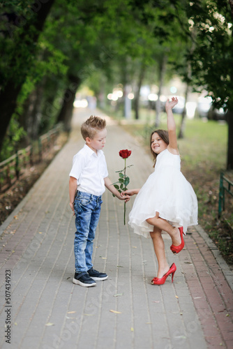 cute couple of children boy and girl, boy gives his girlfriend rose for walk in summer. Girl child in high heels, children play dating and courtship, relationship and growing up concept