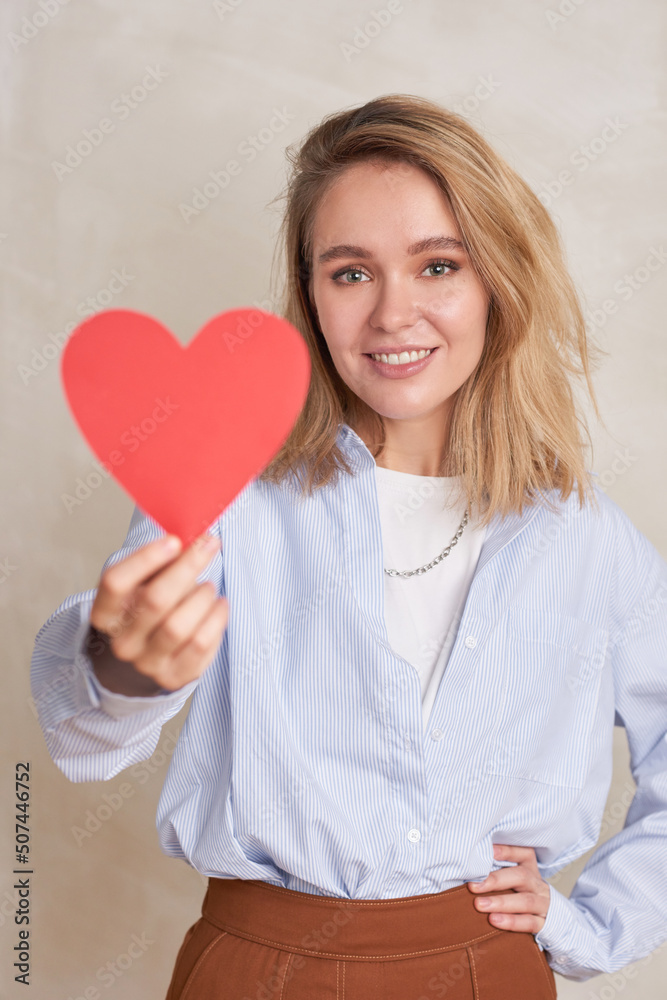 Vertical medium portrait if cheerful young blond woman holding red paper heart smiling at camera Vertical medium portrait if cheerful young blond woman holding red paper heart smiling at camera