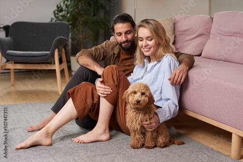 Modern young man and woman in love spending time together at home sitting on floor in living room relaxing with their cute dog
