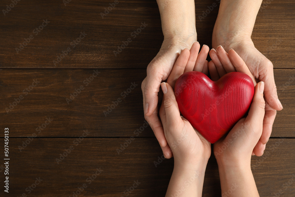 Young and elderly women holding red heart at wooden table, top view. Space for text Young and elderly women holding red heart at wooden table, top view. Space for text