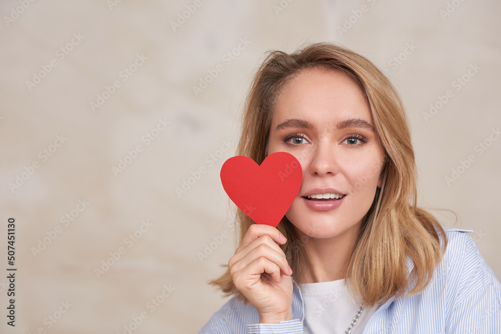 Close-up portrait of attractive young woman in flirty mood holding paper heart looking at camera, copy space
