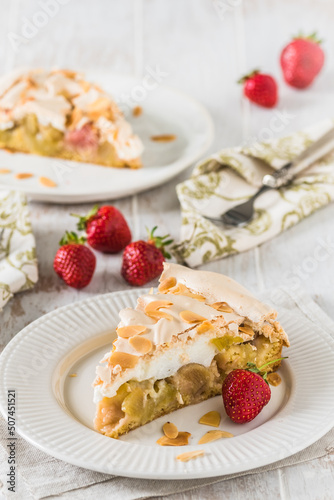 Piece of rhubarb pie with meringue topping and roasted almonds on white wooden background