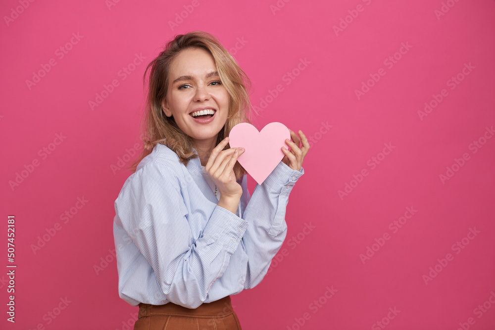 Medium portrait of joyful young woman with blond hair standing against pink wall background holding paper heart smiling at camera Medium portrait of joyful young woman with blond hair standing against pink wall background holding paper heart smiling at camera